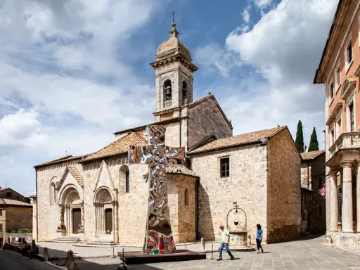 Piazza a San Quirico con una scultura moderna a forma di croce riflettente davanti alla chiesa romanica.