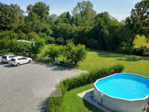 Un **agricampeggio con piscina** immerso nel verde. Auto parcheggiate, camper, biciclette e persone che si rilassano all'ombra degli alberi. La piscina azzurra invita a un tuffo rinfrescante.