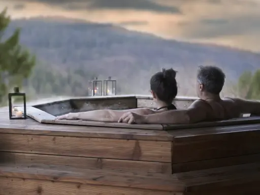 Coppia abbracciata in una vasca idromassaggio di legno, con lanterne accese e vista sulle montagne. Un momento di puro relax in Trentino.