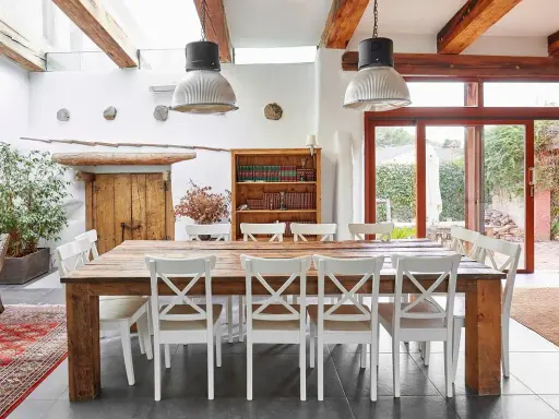 Un rustico tavolo in legno con sedie bianche, illuminato da lampade industriali, in una casa con travi a vista e un giardino che ricorda il Val di Cecina.