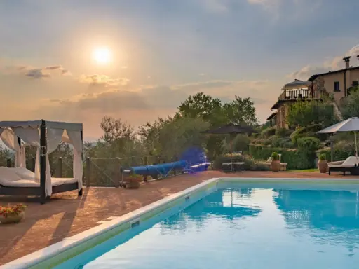 Piscina con lettini e gazebo, ideale per sapere dove dormire in Toscana. Tramonto suggestivo su un casale.