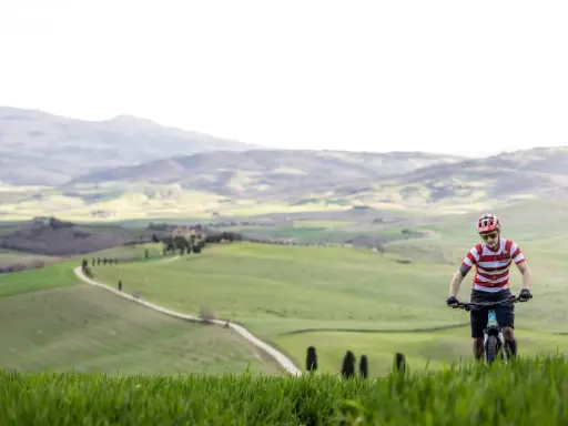 Ciclista in un paesaggio collinare della Toscana, con un sentiero tortuoso che serpeggia tra campi verdi.