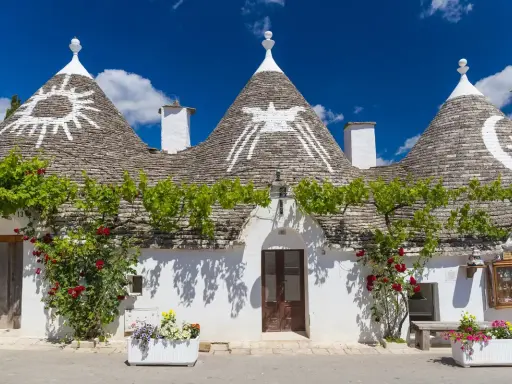 Trulli con simboli sui tetti, circondati da fiori e verde. Un'idea per scoprire **paesi da visitare vicino Alberobello**.