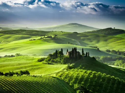 Colline verdi ondulate del Val d'Orcia, con una casa colonica e cipressi su una collina. Un paesaggio da sogno, quasi una mappa visiva della Toscana.