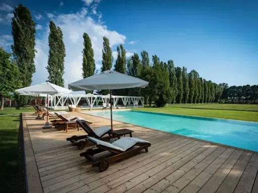 Una splendida piscina in campagna, con lettini prendisole in legno e ombrelloni bianchi, circondata da un prato verde e alti alberi.