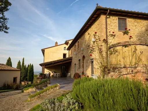 Un casale in pietra con rose rampicanti, un pergolato con tavoli e sedie, e cipressi svettano nel paesaggio della **Toscana in inverno**.