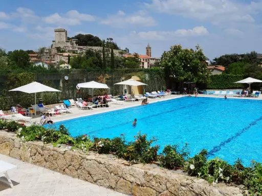 Piscina azzurra in un villaggio famiglie toscana, con castello medievale sullo sfondo e persone che si rilassano al sole.