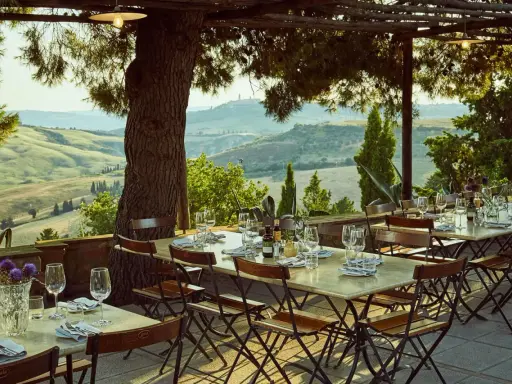 Tavoli apparecchiati sotto un pergolato con vista sulle dolci colline dell'entroterra toscana. Un invito a gustare la cucina locale.