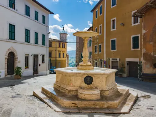 Fontana in pietra nella Valdichiana Senese, circondata da edifici storici con persiane verdi e un cielo azzurro con nuvole bianche.