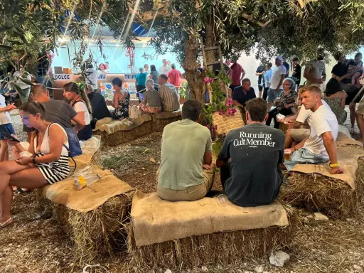 Gente seduta su balle di fieno sotto alberi, un evento estivo in Sicilia.