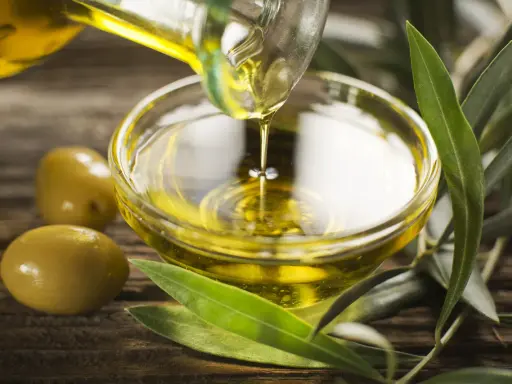 Olio di oliva non extravergine versato in una ciotola di vetro, con olive e foglie d'ulivo su fondo di legno.