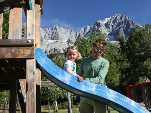 Un padre e una bambina si divertono su uno scivolo in un parco giochi con le maestose montagne della Valle d'Aosta sullo sfondo, un'estate indimenticabile.