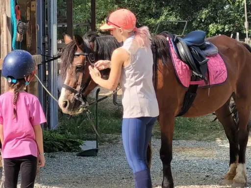 Bambina con casco blu e maglietta rosa osserva un cavallo marrone pronto per un giro a cavallo per bambini. Una ragazza con cappellino arancione prepara il cavallo.