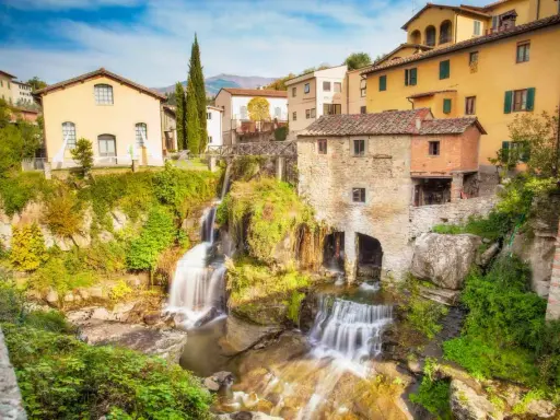 Cascata e mulino in pietra, tra i borghi della Toscana. Case gialle e verdi colline sullo sfondo.
