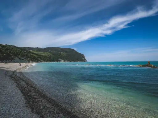 Spiaggia di ciottoli con mare cristallino e una collina verdeggiante, tipica del entroterra marchigiano.