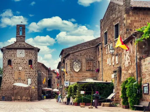 Un vicolo pittoresco a Sovana, Italia, con edifici in pietra, bandiere colorate e un cielo azzurro pieno di nuvole.