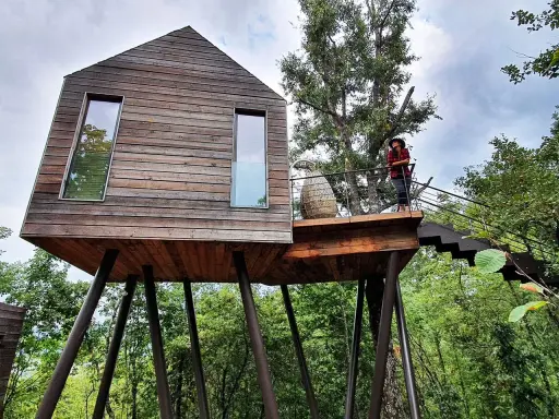 Un'esperienza unica per dormire nella natura toscana: una casa sull'albero in legno, con una persona che ammira il paesaggio dalla terrazza.