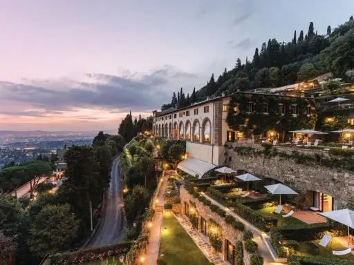 Un lussuoso hotel si affaccia sulle **colline di Firenze** al tramonto, con giardini illuminati e la città in lontananza.