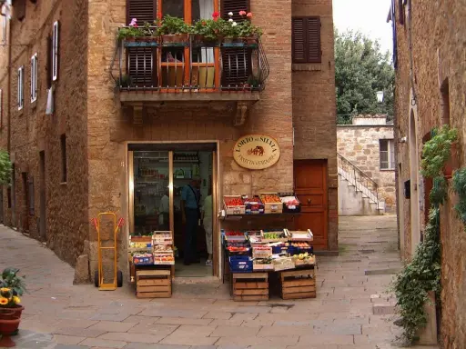 Un piccolo negozio di frutta e verdura in un vicolo di **paesi toscani**. Frutta fresca in cassette di legno, un balcone fiorito e l'atmosfera autentica.