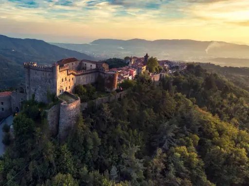 Antico castello medievale su una collina boscosa, uno dei tanti castelli da visitare in Toscana, con un villaggio arroccato e vista sulla valle al tramonto.