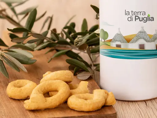 Taralli pugliesi su tagliere di legno, accanto a un contenitore con il logo "la terra di Puglia" e un ramo d'ulivo.