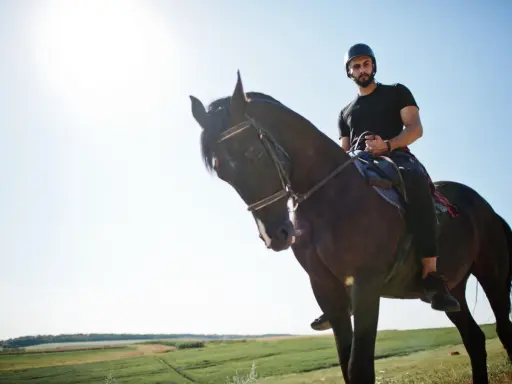 Uomo con casco e barba cavalca un cavallo scuro in un paesaggio verdeggiante. Un'esperienza di **Toscana a cavallo**.