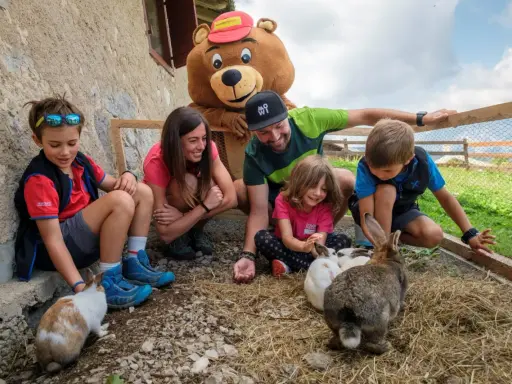 Bambini e adulti interagiscono con conigli in un recinto, parte di un'esperienza in fattorie didattiche. Un personaggio con costume da orso sorride.