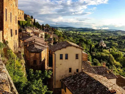 Panoramica di borghi toscana in treno, con case in pietra e tegole rosse su colline verdi sotto un cielo nuvoloso.