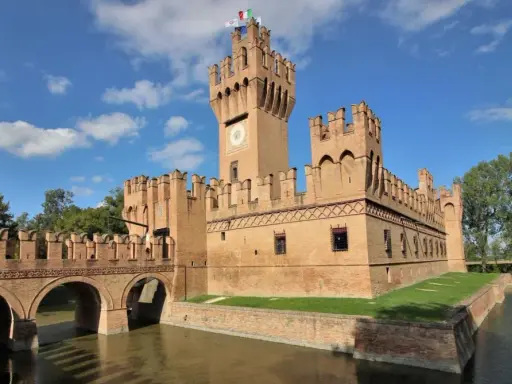 Un imponente castello in mattoni rossi, uno dei tanti castelli Emilia Romagna, con un ponte ad archi sull'acqua e torri merlate sotto un cielo azzurro.