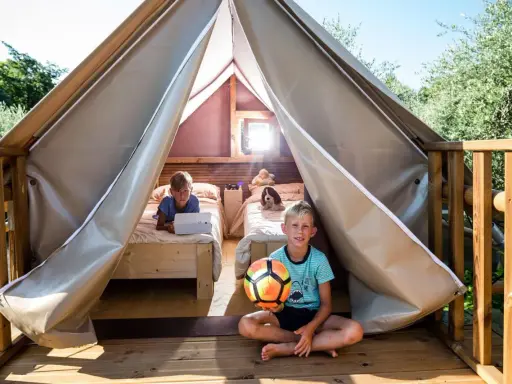 Bambini felici in un glamping: uno gioca col pallone, l'altro usa un tablet a letto. Avventura e relax per i più piccoli.