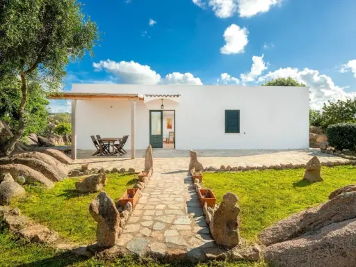 Un sentiero in pietra conduce a un bianco casolare, circondato da rocce granitiche e vegetazione mediterranea. Ideale per scoprire gli agriturismi Sardegna nord.