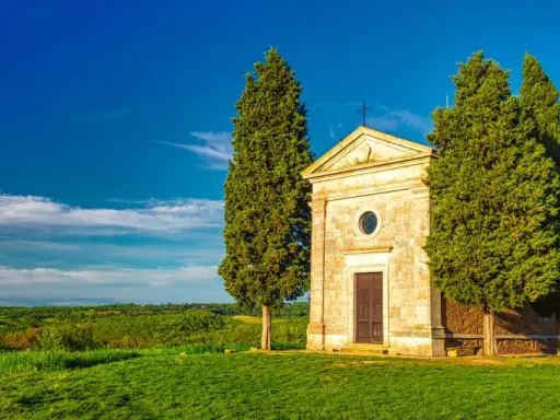 Cappella di San Quirico d'Orcia, circondata da cipressi, su un prato verde sotto un cielo azzurro.