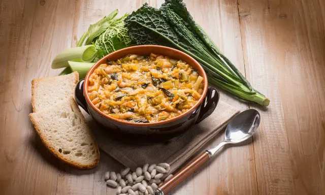 Piatto tipico toscano con pane raffermo: ribollita fumante in ciotola, contornata da fagioli, cavolo nero e porri.