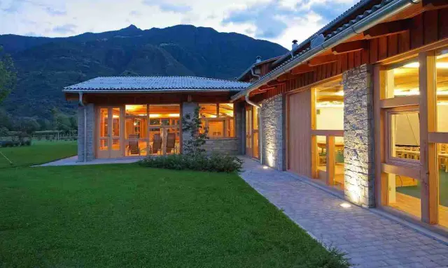 Edificio moderno con pietra e legno, circondato da prati verdi, con ampie vetrate che illuminano l'interno. Sembra un luogo ideale per fattorie con ristorante.