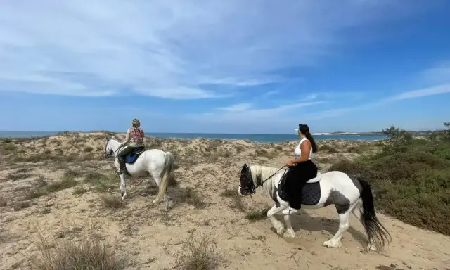 Due donne godono di una passeggiata a cavallo in Sicilia, tra dune sabbiose e il mare azzurro.