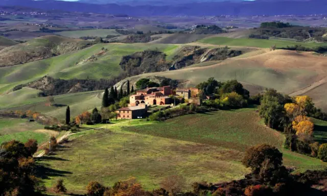 Colline ondulate e campi coltivati, un casale in mattoni rossi e cipressi svettanti, tipici delle **strade panoramiche toscana**.