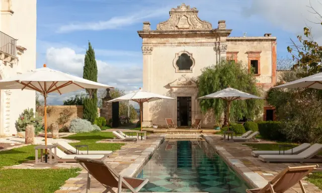 Piscina e lettini in una masseria per bambini in Puglia, con un antico edificio sullo sfondo.