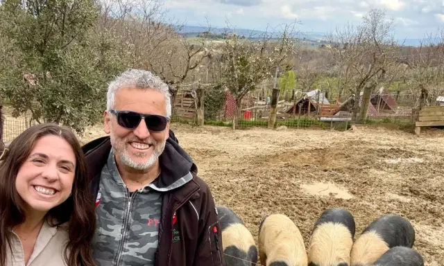 Tre amici sorridono mentre salutano i maiali senesi in un recinto fangoso, con un paesaggio collinare sullo sfondo.