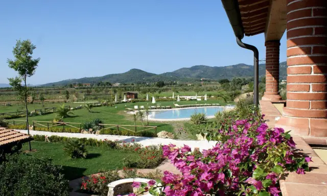 Un angolo di campagna toscana lungo il mare, con piscina, giardini curati e colline verdi all'orizzonte.