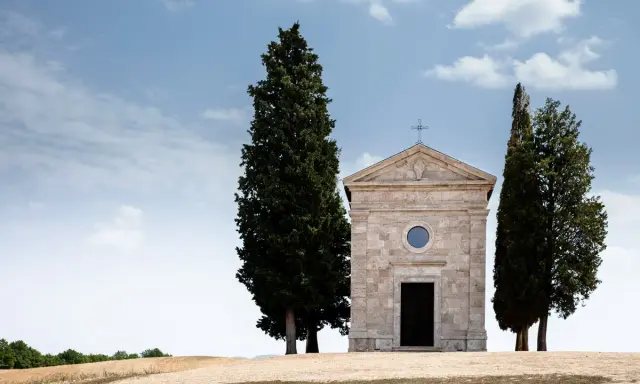 La cappella della Madonna di Vitaleta, con i suoi cipressi iconici, si erge solitaria su una collina toscana sotto un cielo azzurro e nuvoloso.