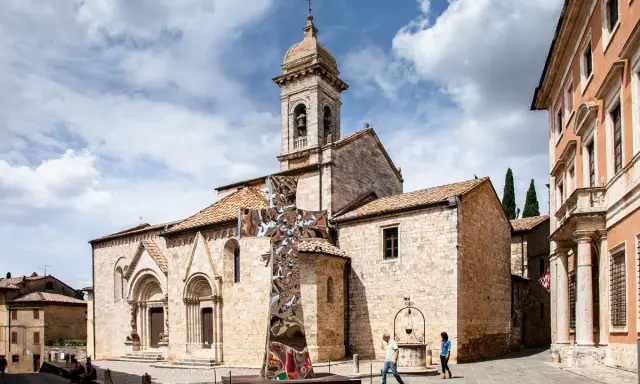 Piazza a San Quirico con una scultura moderna a forma di croce riflettente davanti alla chiesa romanica.