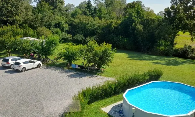 Un **agricampeggio con piscina** immerso nel verde. Auto parcheggiate, camper, biciclette e persone che si rilassano all'ombra degli alberi. La piscina azzurra invita a un tuffo rinfrescante.