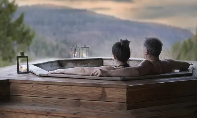 Coppia abbracciata in una vasca idromassaggio di legno, con lanterne accese e vista sulle montagne. Un momento di puro relax in Trentino.