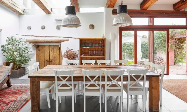 Un rustico tavolo in legno con sedie bianche, illuminato da lampade industriali, in una casa con travi a vista e un giardino che ricorda il Val di Cecina.
