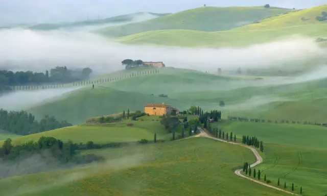 Dolci colline verdi avvolte dalla nebbia mattutina nel Val di Norcia, con casali in pietra e filari di cipressi.