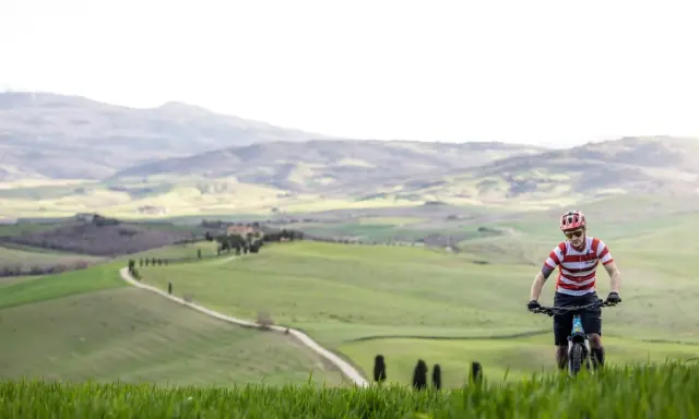 Ciclista in un paesaggio collinare della Toscana, con un sentiero tortuoso che serpeggia tra campi verdi.