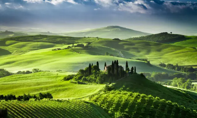 Colline verdi ondulate del Val d'Orcia, con una casa colonica e cipressi su una collina. Un paesaggio da sogno, quasi una mappa visiva della Toscana.