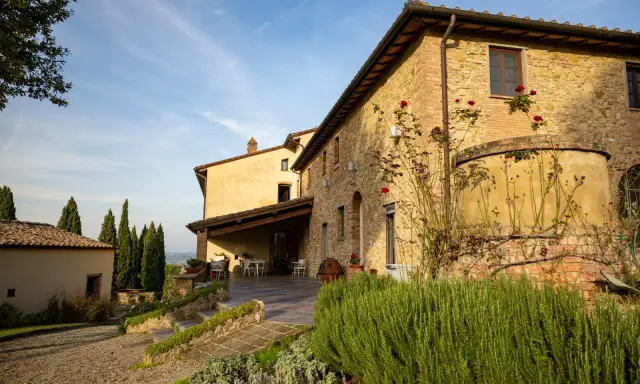Un casale in pietra con rose rampicanti, un pergolato con tavoli e sedie, e cipressi svettano nel paesaggio della **Toscana in inverno**.