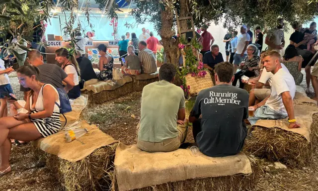 Gente seduta su balle di fieno sotto alberi, un evento estivo in Sicilia.