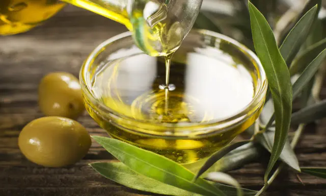 Olio di oliva non extravergine versato in una ciotola di vetro, con olive e foglie d'ulivo su fondo di legno.